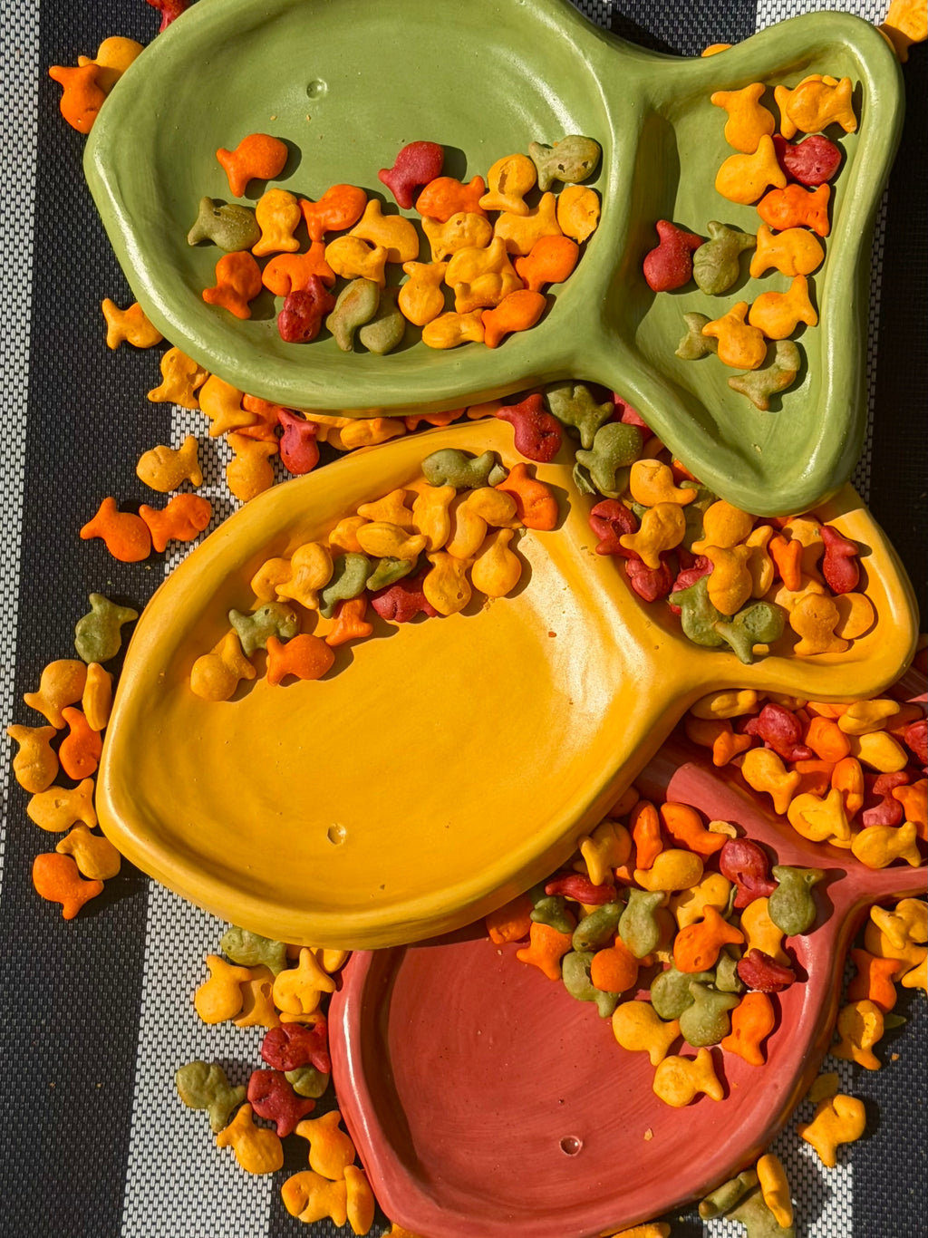 Three ceramic fish-shaped pet bowls in green, yellow, and terracotta, filled with colorful kibble, arranged on a black and white checkered mat.