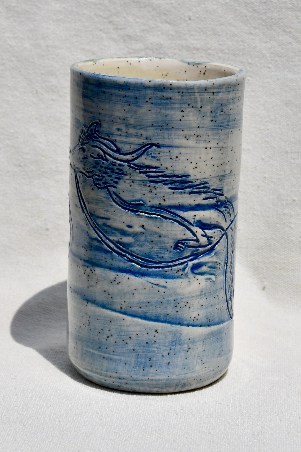 A handmade ceramic cup with a blue and white speckled glaze, featuring a blue painted bird design on the side, set against a white textured background.