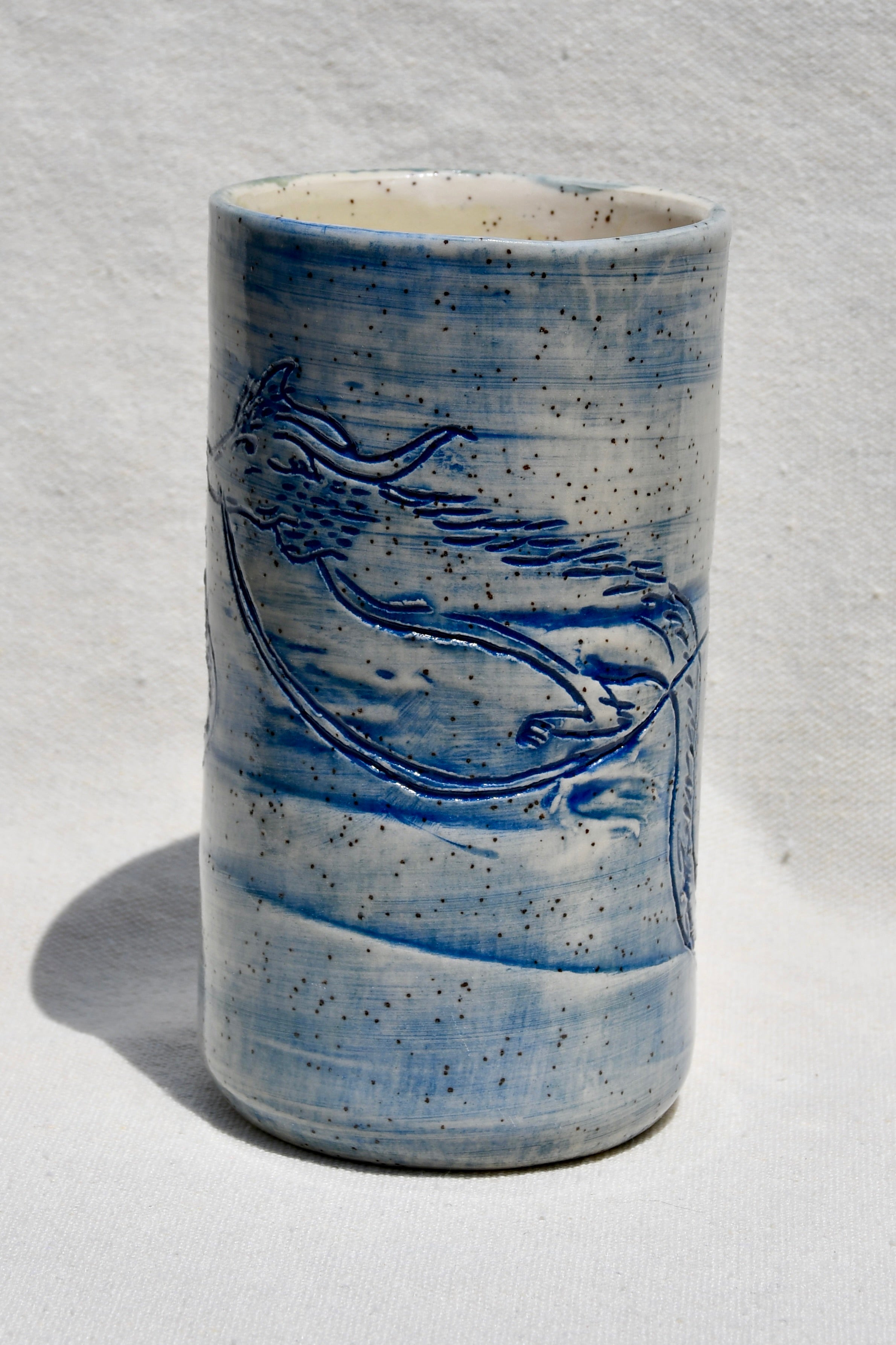 A handmade ceramic cup with a blue and white speckled glaze, featuring a blue painted bird design on the side, set against a white textured background.