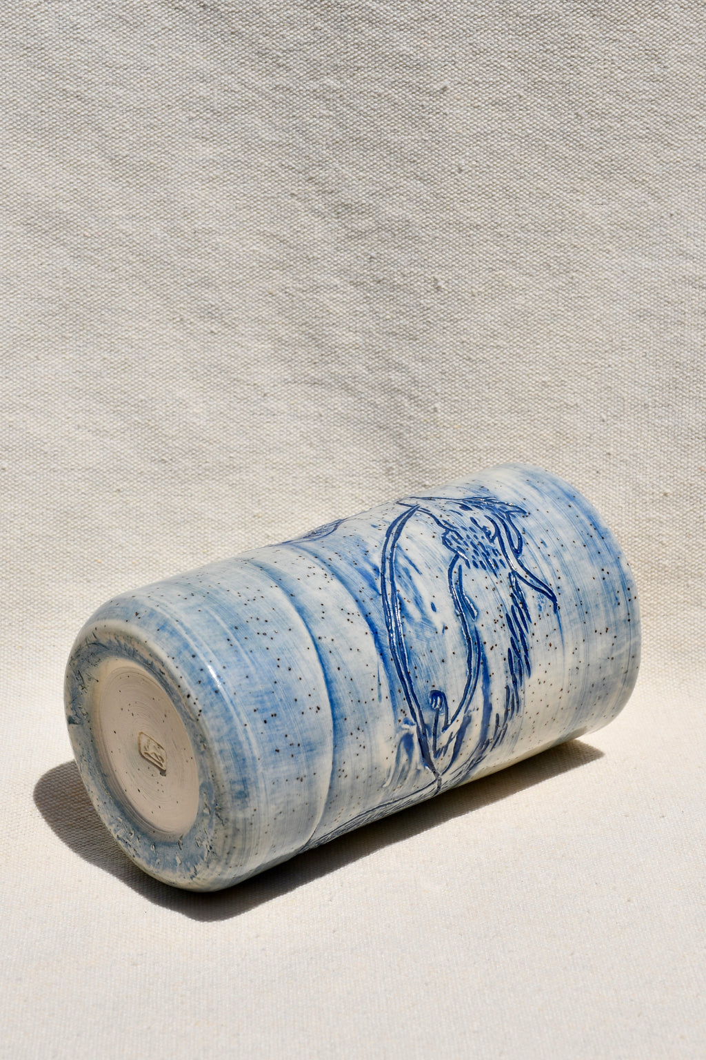 A handmade ceramic cylinder vase with a blue and white speckled glaze and a blue floral design, lying on its side against a textured off-white background.