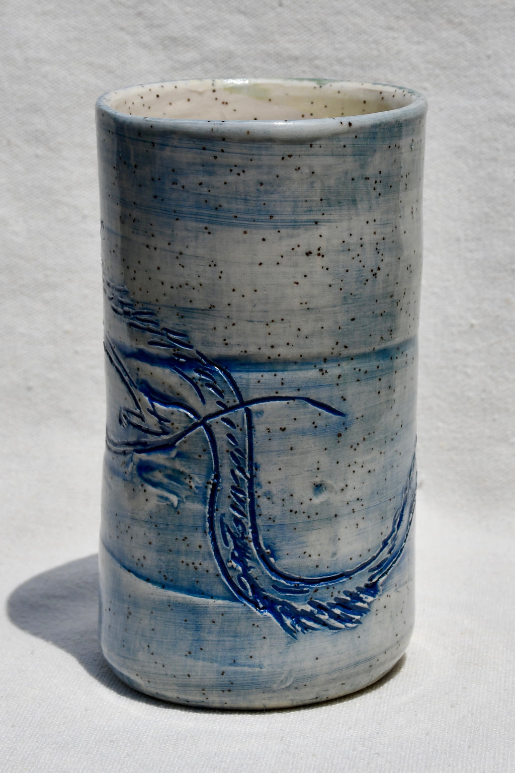 A handmade ceramic cup with a cylindrical shape, featuring a speckled light blue and white glaze with a blue painted dragon design on the front, set against a white textured background.