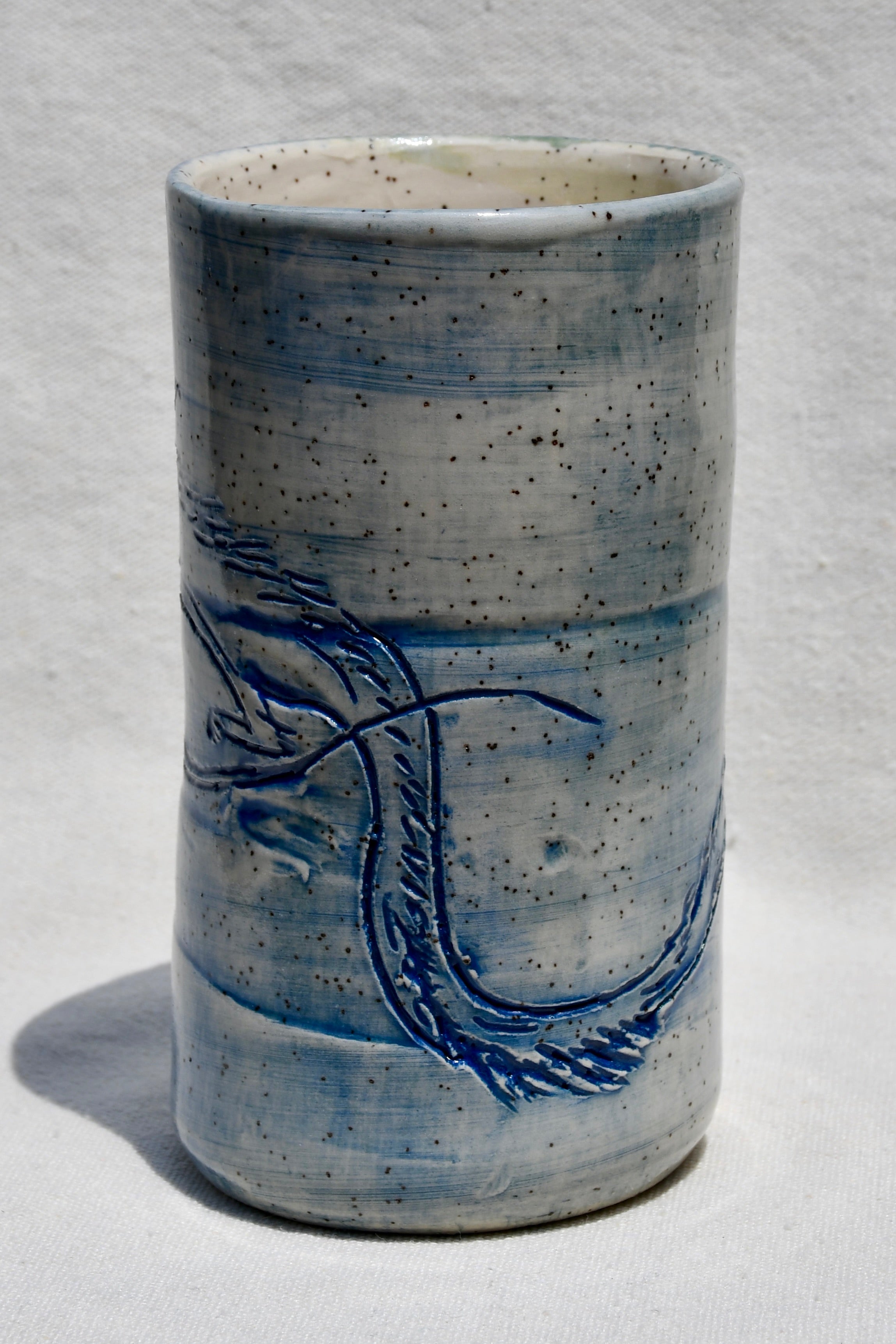 A handmade ceramic cup with a cylindrical shape, featuring a speckled light blue and white glaze with a blue painted dragon design on the front, set against a white textured background.