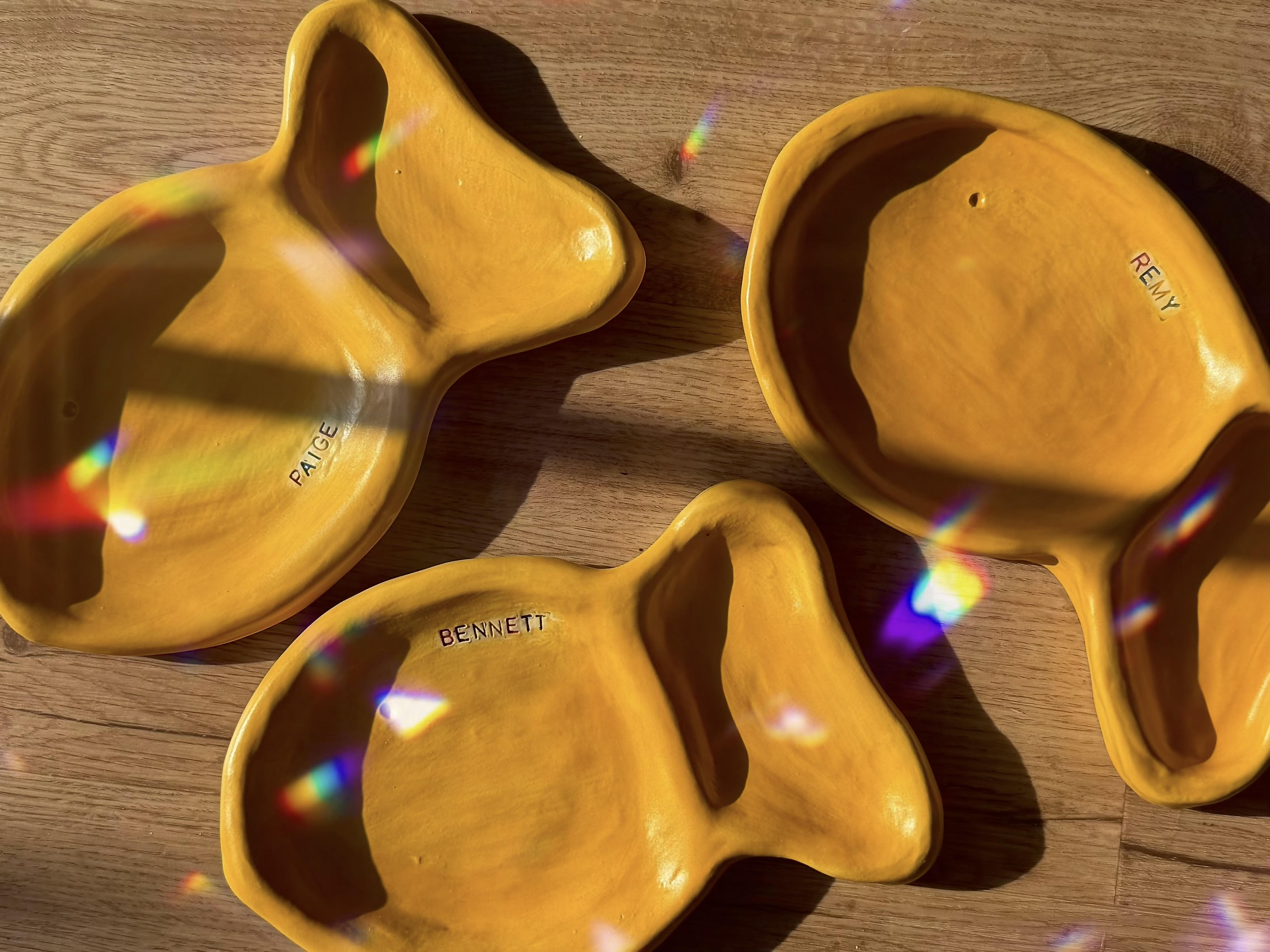 Three yellow ceramic goldfish plates with 'Bennett' and 'Remy' branding on a wooden surface.